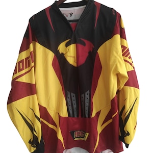 May include: A long-sleeved jersey in yellow, black, and maroon, hanging on a wooden hanger. The jersey features a V-neck and a design with geometric shapes and the number "1997" on the front. The jersey has a sporty aesthetic.