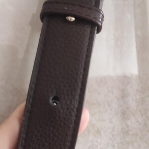 May include: Close-up of a dark brown leather strap with a textured surface. The strap has a buckle and a series of holes. The strap is likely part of a bag or belt.