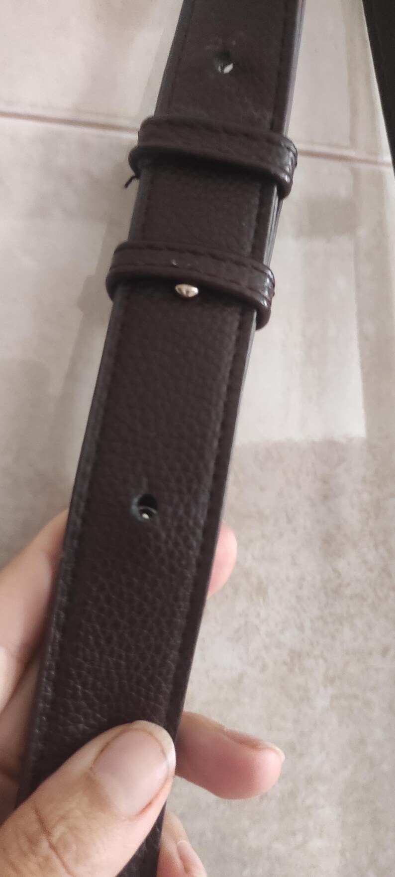 May include: Close-up of a dark brown leather strap with a textured surface. The strap has a buckle and a series of holes. The strap is likely part of a bag or belt.