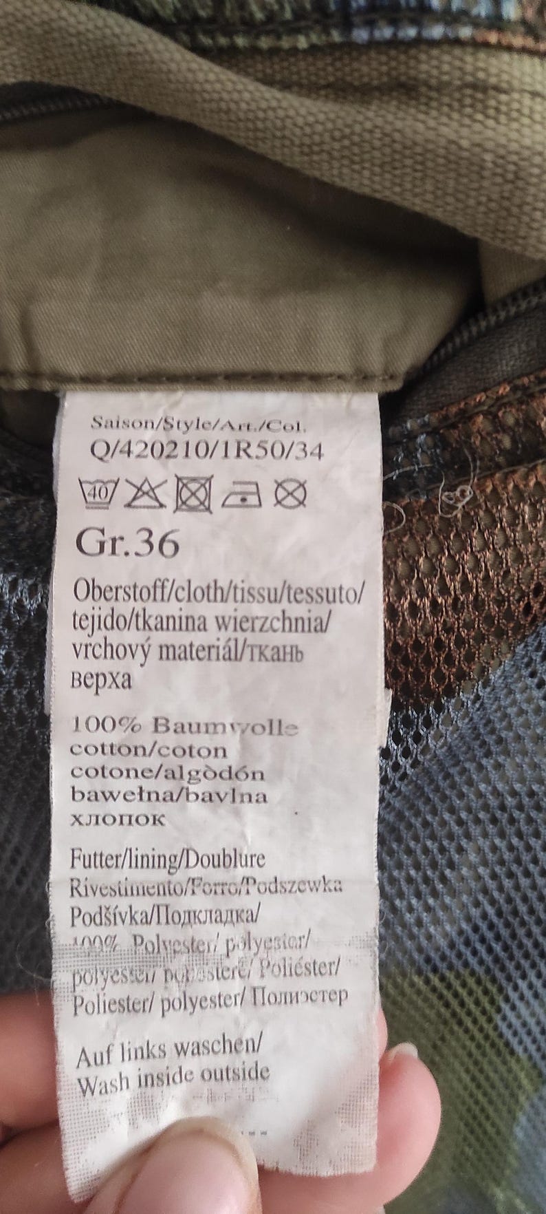 May include: A white clothing label with black text and care instructions. The label says "Saison/Style/Art./Col. Q/420210/1R50/34" and "Gr. 36". The label also includes instructions for washing and ironing the garment.