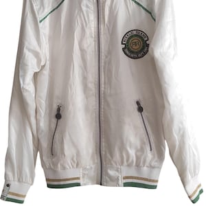 May include: White zip-up jacket with green and gold trim. The jacket features a ribbed collar, cuffs, and hem with stripes. A circular patch on the chest reads "RIVALDI BRAND SINCE SEPT 1984".