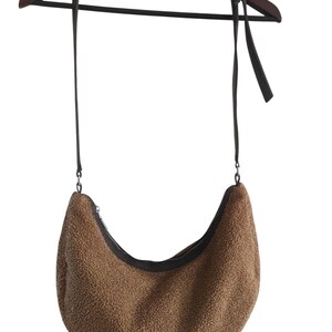 May include: A brown, crescent-shaped shoulder bag with a textured, fuzzy exterior. The bag features a black zipper and a long, dark brown strap. The bag is hanging on a wooden hanger.