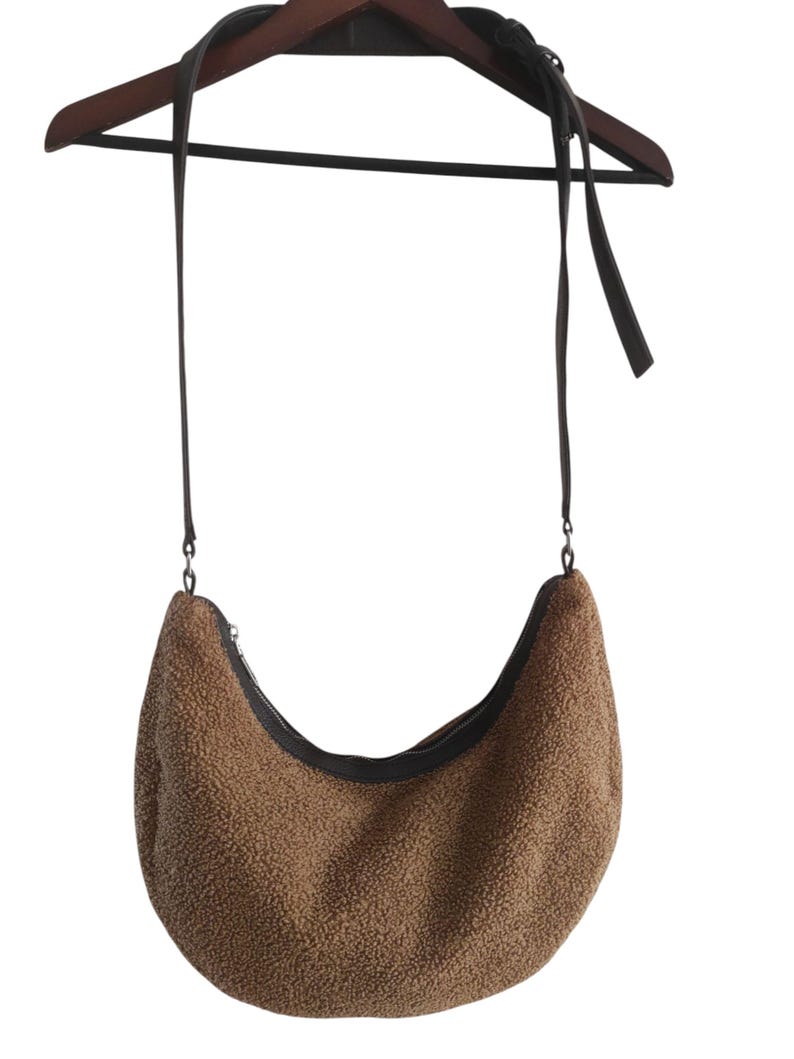 May include: A brown, crescent-shaped shoulder bag with a textured, fuzzy exterior. The bag features a black zipper and a long, dark brown strap. The bag is hanging on a wooden hanger.