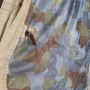 May include: A beige jacket with a camouflage pattern lining. The lining is a mesh fabric with a brown, green, and tan camouflage pattern. The jacket has a pocket on the inside with a black button.