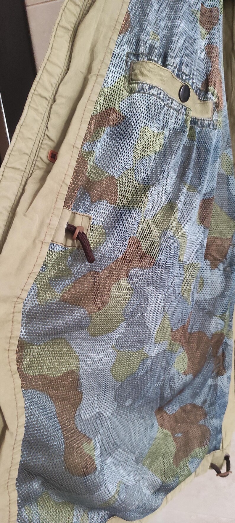 May include: A beige jacket with a camouflage pattern lining. The lining is a mesh fabric with a brown, green, and tan camouflage pattern. The jacket has a pocket on the inside with a black button.