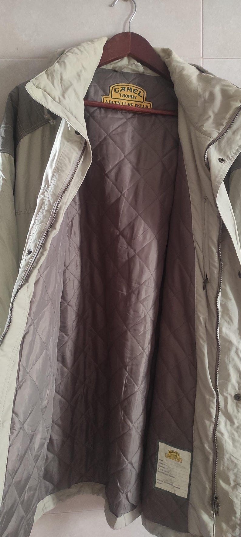 May include: A beige and brown jacket with a quilted lining. The jacket has a zipper closure and a label that reads "Camel Trophy Adventure Wear".