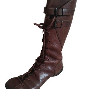 May include: A brown leather boot with a lace-up front and two buckle straps. The boot has a rounded toe and a tall shaft, and is made of textured leather. The boot is designed to be worn on one foot.