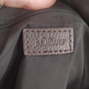 May include: Close-up of a brown leather tag with the embossed text "s.Oliver" stitched onto a dark brown bag. The tag is rectangular with rounded corners and a textured surface.
