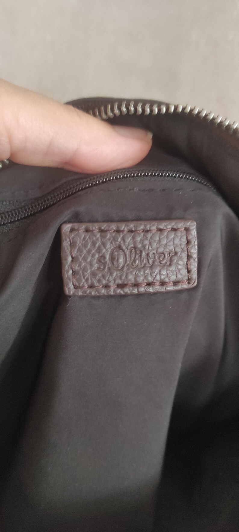 May include: Close-up of a brown leather tag with the embossed text "s.Oliver" stitched onto a dark brown bag. The tag is rectangular with rounded corners and a textured surface.
