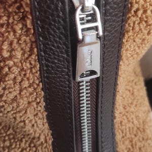 May include: Close-up of a brown and tan textured bag. The bag features a dark brown leather trim and a silver zipper. The zipper pull is rectangular with the brand name engraved on it.