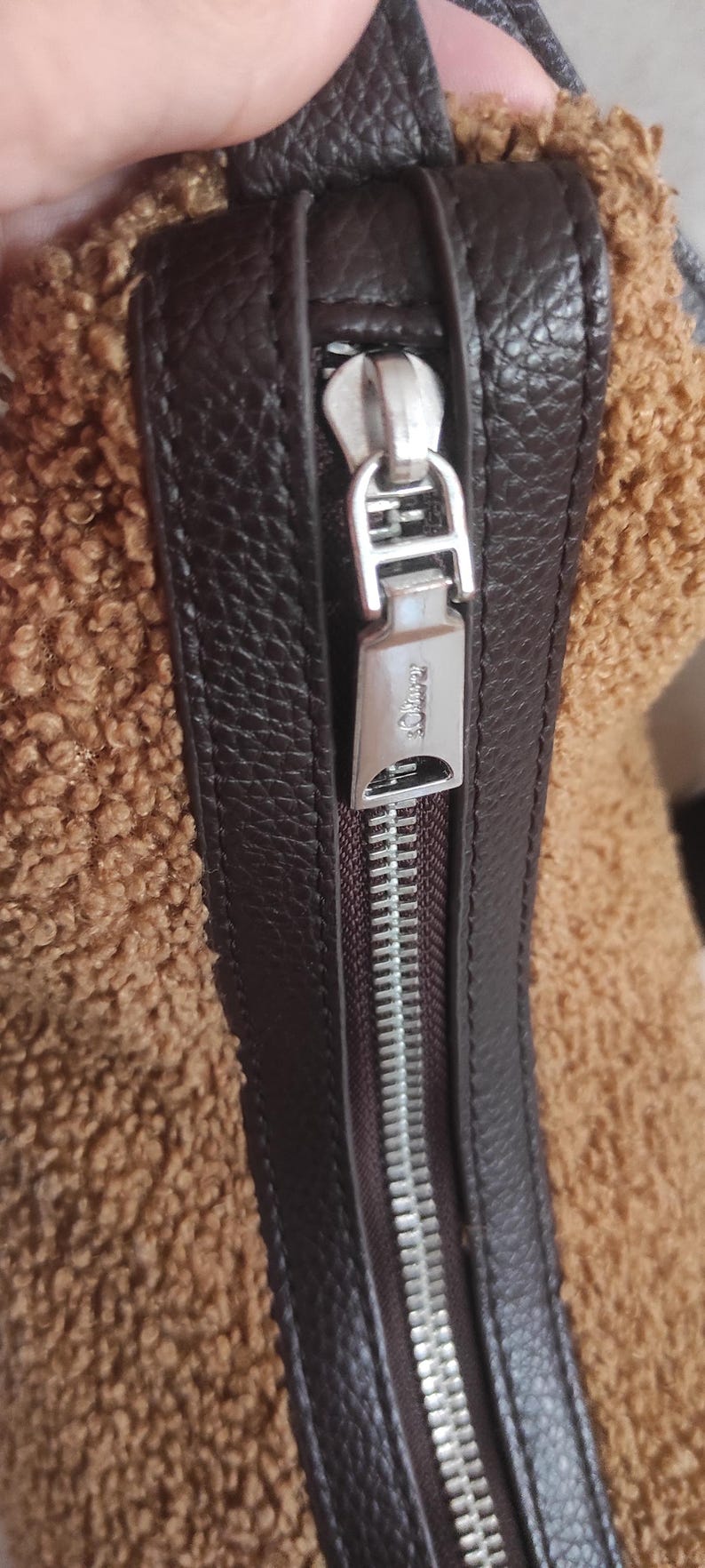 May include: Close-up of a brown and tan textured bag. The bag features a dark brown leather trim and a silver zipper. The zipper pull is rectangular with the brand name engraved on it.