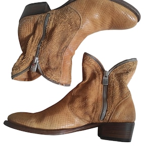 May include: Two tan ankle boots with a textured, snakeskin-like pattern. Each boot features a side zipper with a silver-colored pull. The boots have a low heel and a rounded toe.