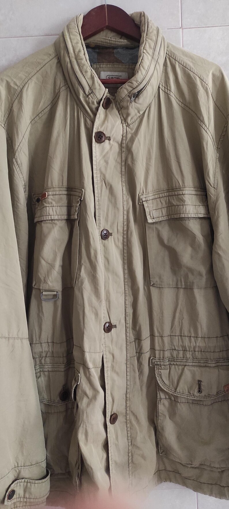 May include: A beige utility jacket with multiple pockets and a button closure. The jacket has a brand label that reads "Camel" on the front.