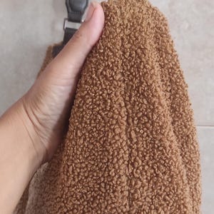 May include: A brown, fluffy teddy bear-like bag with a dark brown strap and silver hardware. The bag is held by a hand, showcasing the soft, textured material. The bag's design is simple and casual.
