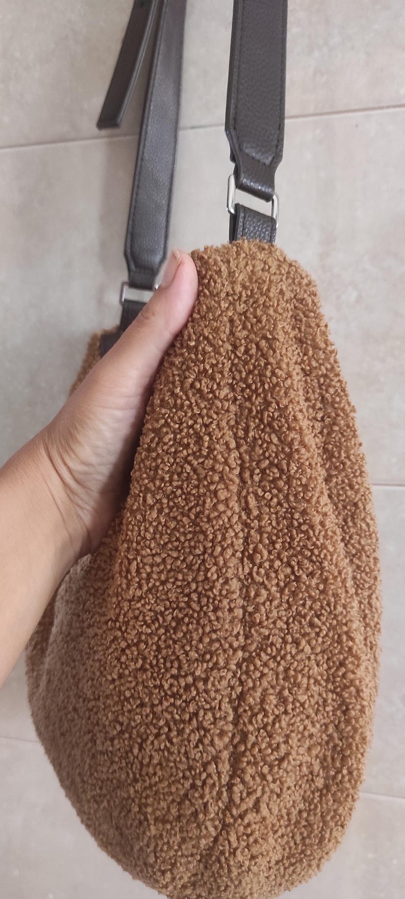 May include: A brown, fluffy teddy bear-like bag with a dark brown strap and silver hardware. The bag is held by a hand, showcasing the soft, textured material. The bag's design is simple and casual.