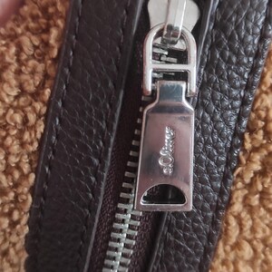 May include: Close-up of a brown handbag with a textured leather strap and a silver zipper. The zipper pull has the brand name "s.Oliver" engraved on it. The bag has a fuzzy, tan-colored material.