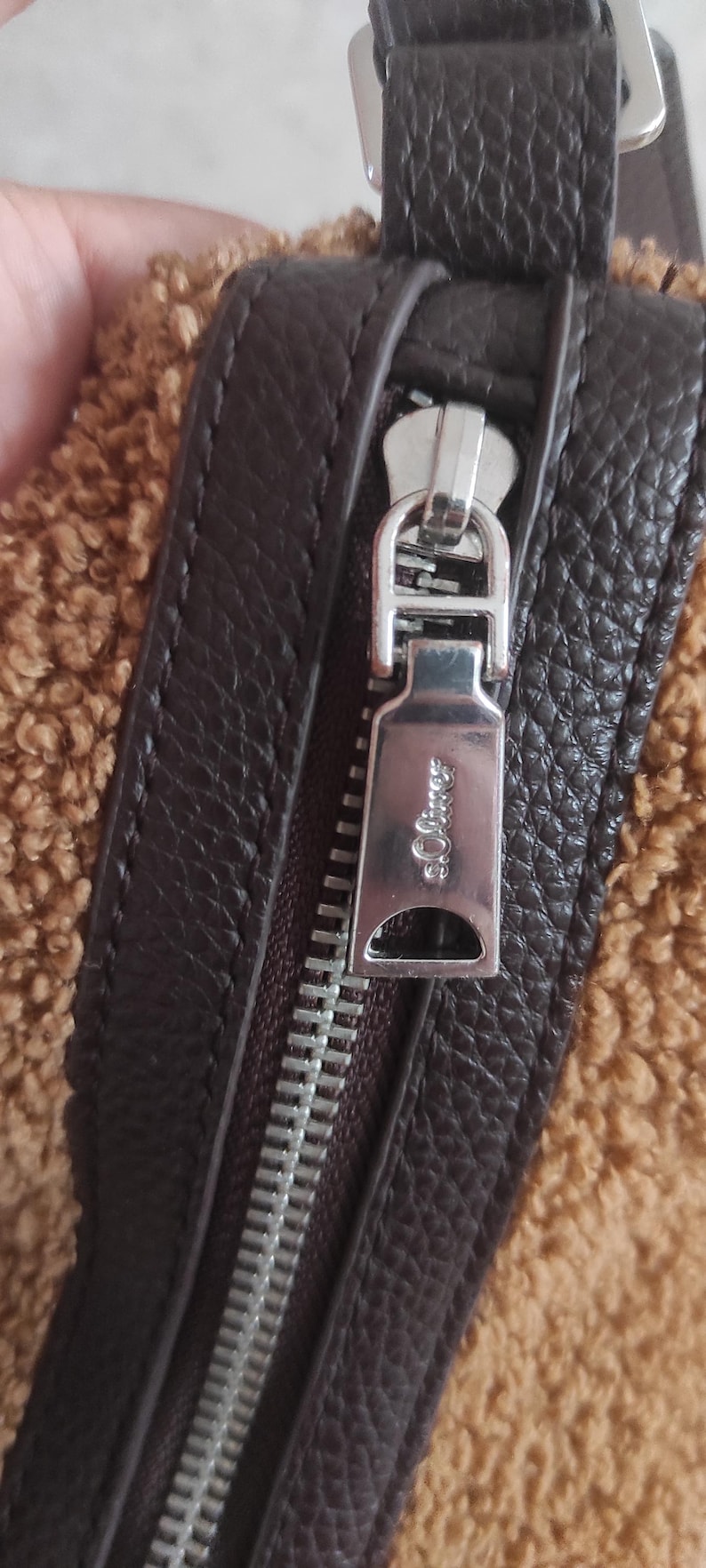 May include: Close-up of a brown handbag with a textured leather strap and a silver zipper. The zipper pull has the brand name "s.Oliver" engraved on it. The bag has a fuzzy, tan-colored material.