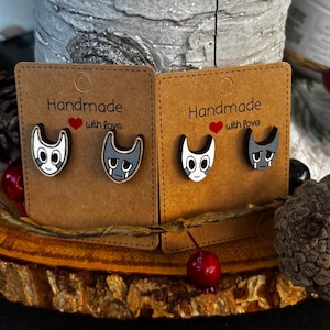 Puede incluir: Dos pares de pendientes de bot&oacute;n hechos a mano con forma de gato. Un par presenta una cara de gato blanca con detalles negros, el otro par tiene una cara de gato gris con l&aacute;grimas. Cada par est&aacute; montado en una tarjeta marr&oacute;n con el texto "Handmade with love".
