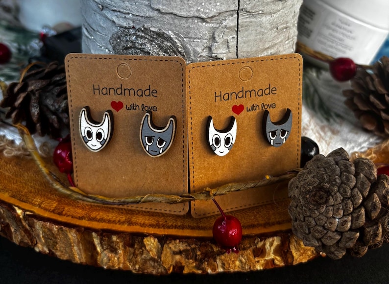 Puede incluir: Dos pares de pendientes de bot&oacute;n hechos a mano con forma de gato. Un par presenta una cara de gato blanca con detalles negros, el otro par tiene una cara de gato gris con l&aacute;grimas. Cada par est&aacute; montado en una tarjeta marr&oacute;n con el texto "Handmade with love".