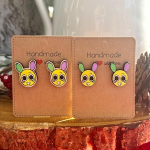Puede incluir: Dos pares de pendientes hechos a mano con caras de conejito en tarjetas marrones. Los pendientes presentan caras amarillas con ojos y bocas negras, y orejas coloridas. Las tarjetas están etiquetadas como "Handmade with Love."