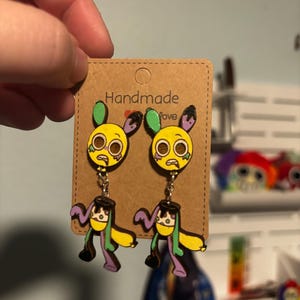 Puede incluir: Un par de pendientes hechos a mano con un dise&ntilde;o de personaje de dibujos animados amarillo, con detalles verdes, morados y negros. Los pendientes est&aacute;n sujetos a una tarjeta marr&oacute;n con las palabras "Handmade Love".