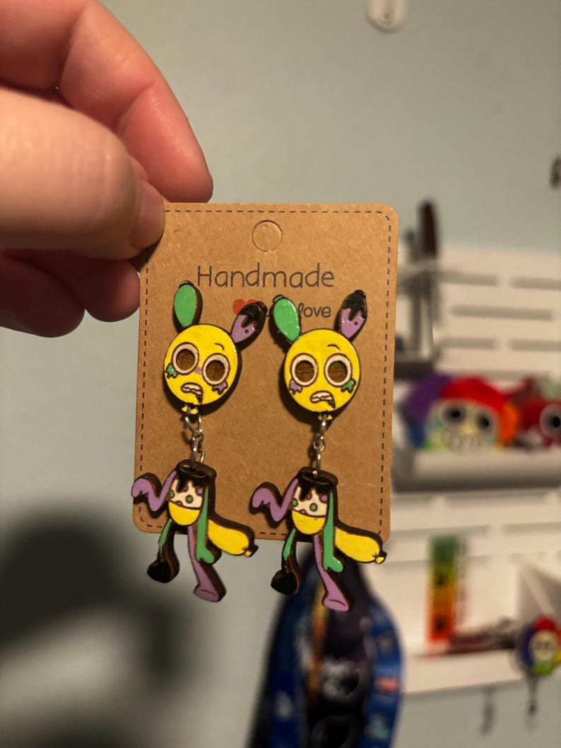 Puede incluir: Un par de pendientes hechos a mano con un dise&ntilde;o de personaje de dibujos animados amarillo, con detalles verdes, morados y negros. Los pendientes est&aacute;n sujetos a una tarjeta marr&oacute;n con las palabras "Handmade Love".