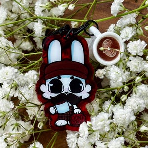 May include: A keychain featuring a cartoon rabbit with a mustache wearing a brown cap and holding a blue mug, with a small white coffee cup charm attached. The rabbit has large black eyes and is set against a translucent red acrylic background.