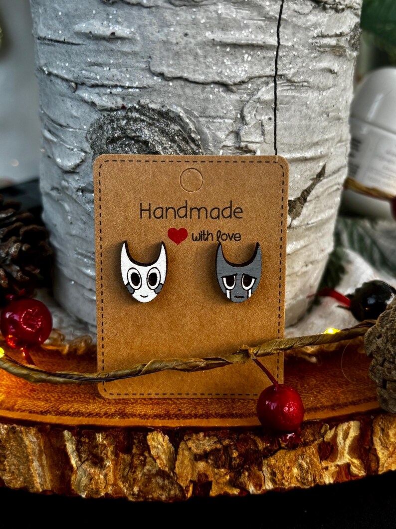 Puede incluir: Pendientes de madera con dos caras de dibujos animados, una feliz y otra triste, presentados en una tarjeta marr&oacute;n. La tarjeta dice "Handmade with love".