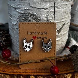 Puede incluir: Pendientes de bot&oacute;n de madera con caras de gato de dibujos animados, uno sonriendo y otro triste, sobre una tarjeta marr&oacute;n. La tarjeta dice "Handmade with love" en texto negro con un coraz&oacute;n rojo. Los pendientes se exhiben sobre una superficie de madera.