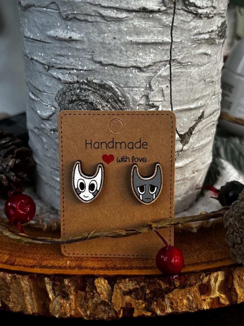 Puede incluir: Pendientes de bot&oacute;n de madera con caras de gato de dibujos animados, uno sonriendo y otro triste, sobre una tarjeta marr&oacute;n. La tarjeta dice "Handmade with love" en texto negro con un coraz&oacute;n rojo. Los pendientes se exhiben sobre una superficie de madera.