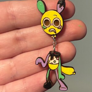 Puede incluir: Un pendiente de madera de estilo caricatura, que representa una cara amarilla con ojos grandes y una expresi&oacute;n triste, conectada por una cadena a un cuerpo con un dise&ntilde;o amarillo, morado y verde. El pendiente tiene una est&eacute;tica &uacute;nica y caprichosa.