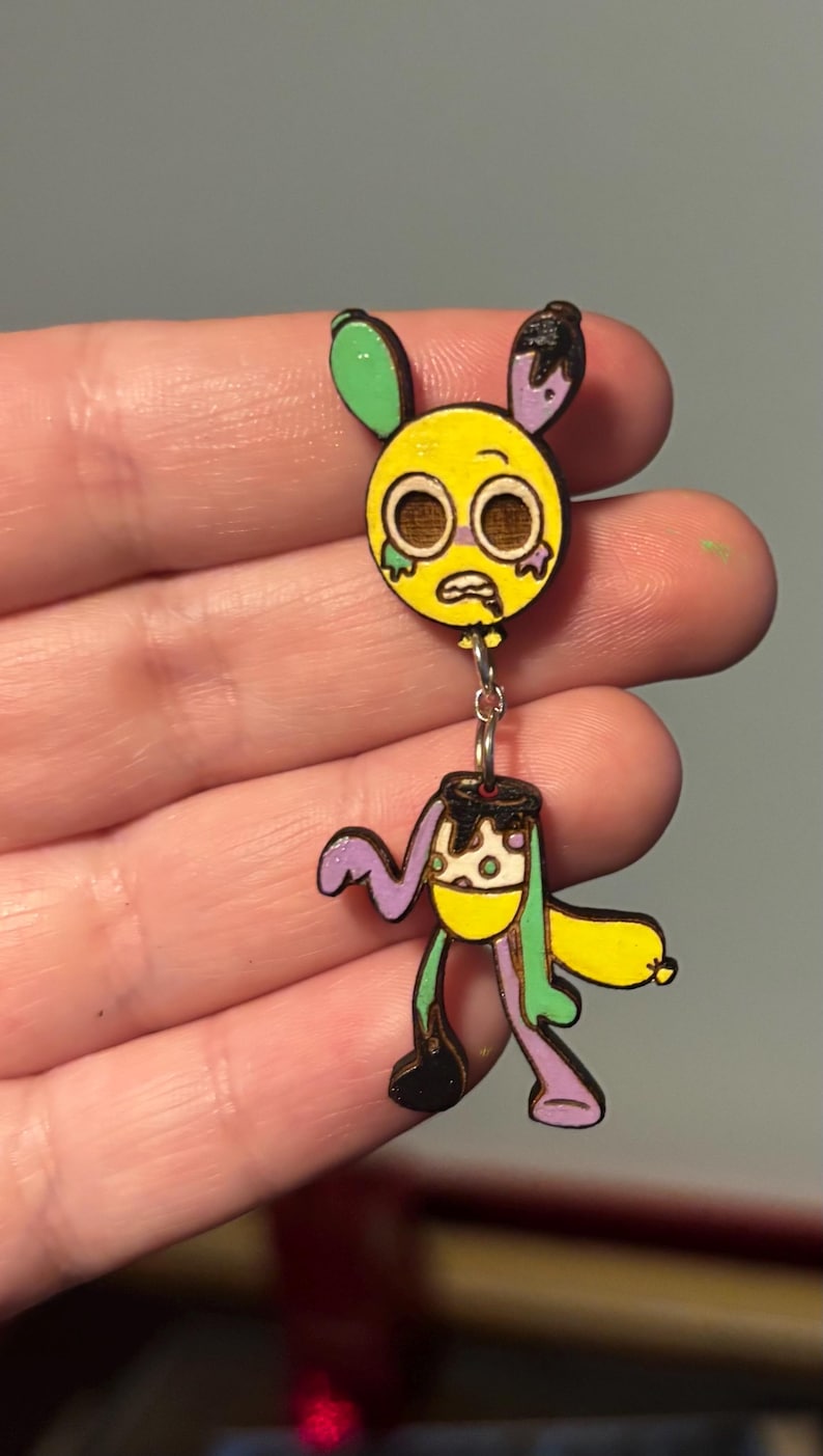 Puede incluir: Un pendiente de madera de estilo caricatura, que representa una cara amarilla con ojos grandes y una expresi&oacute;n triste, conectada por una cadena a un cuerpo con un dise&ntilde;o amarillo, morado y verde. El pendiente tiene una est&eacute;tica &uacute;nica y caprichosa.