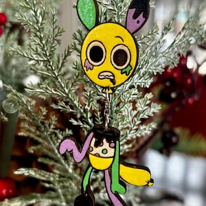 Puede incluir: Adorno de madera con un personaje de dibujos animados con cabeza amarilla, ojos negros y expresión angustiada. El personaje tiene detalles en morado y verde, suspendido por una cadena plateada. El adorno está sobre un árbol de Navidad.