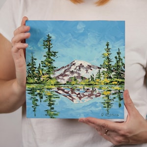 May include: Square oil painting depicting a mountain landscape with a snow-covered peak reflected in the water. The sky is blue with white brushstrokes, and the trees are green. Signed by O. Rodina.