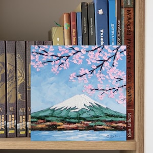 May include: Square painting depicting Mount Fuji with a snow-covered peak, green slopes, and a blue lake. Pink cherry blossom branches are in the foreground against a light blue sky. Signed by O. Rodion.