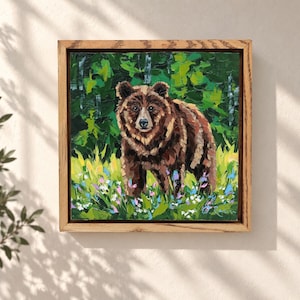 Op de afbeelding: Een vierkant olieverfschilderij in een lichte houten lijst met een bruine beer die in een veld met groen gras en kleurrijke wilde bloemen staat. De achtergrond toont een dicht bos van groene bomen met dikke, zichtbare penseelstreken.
