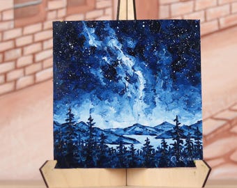 May include: An original acrylic painting on canvas depicting a night sky with a starry galaxy and a landscape of mountains and trees. The artwork is displayed on a small wooden easel.