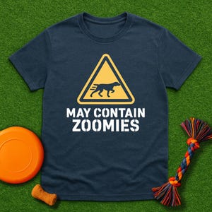 Puede incluir: Camiseta azul marino con un letrero de advertencia amarillo con la silueta de un perro y el texto "MAY CONTAIN ZOOMIES" en blanco. También se ven un frisbee naranja, un pequeño juguete para masticar y un juguete de cuerda.
