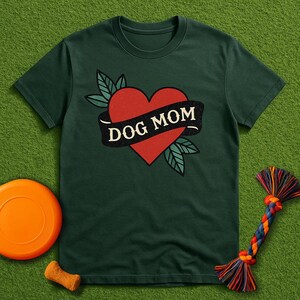 Könnte beinhalten: Dunkelgrünes T-Shirt mit einem roten Herz und einem schwarzen Banner mit der Aufschrift "DOG MOM". Das Herz ist von grünen Blättern umgeben. Eine orangefarbene Frisbeescheibe, ein Hundekeks und ein farbenfrohes Seilspielzeug sind ebenfalls zu sehen.
