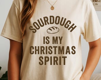 May include: Cream-colored t-shirt with the text "SOURDOUGH IS MY CHRISTMAS SPIRIT" in brown lettering. The word "SOURDOUGH" arches above a bread loaf illustration. The shirt has short sleeves and a relaxed fit, suitable for casual wear.