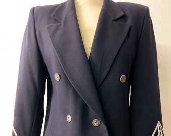Könnte beinhalten: Marineblauer, doppelreihiger Blazer mit silberfarbenen Knöpfen. Der Blazer hat dekorative silberne Stickereien an den Manschetten und Oberärmeln. Eine klassische, maßgeschneiderte Jacke.