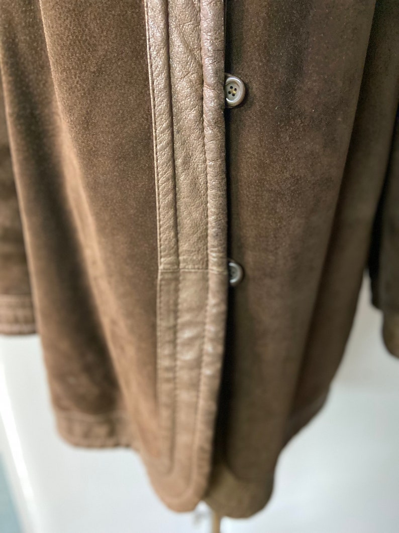 May include: Close-up of a brown suede jacket with a brown leather trim and two buttons. The jacket has a classic design and a soft, luxurious feel.