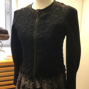 Puede incluir: Chaqueta negra con cierre de cremallera y detalles de encaje intrincados. La chaqueta tiene un dobladillo con volantes y est&aacute; hecha de una tela suave y texturizada.