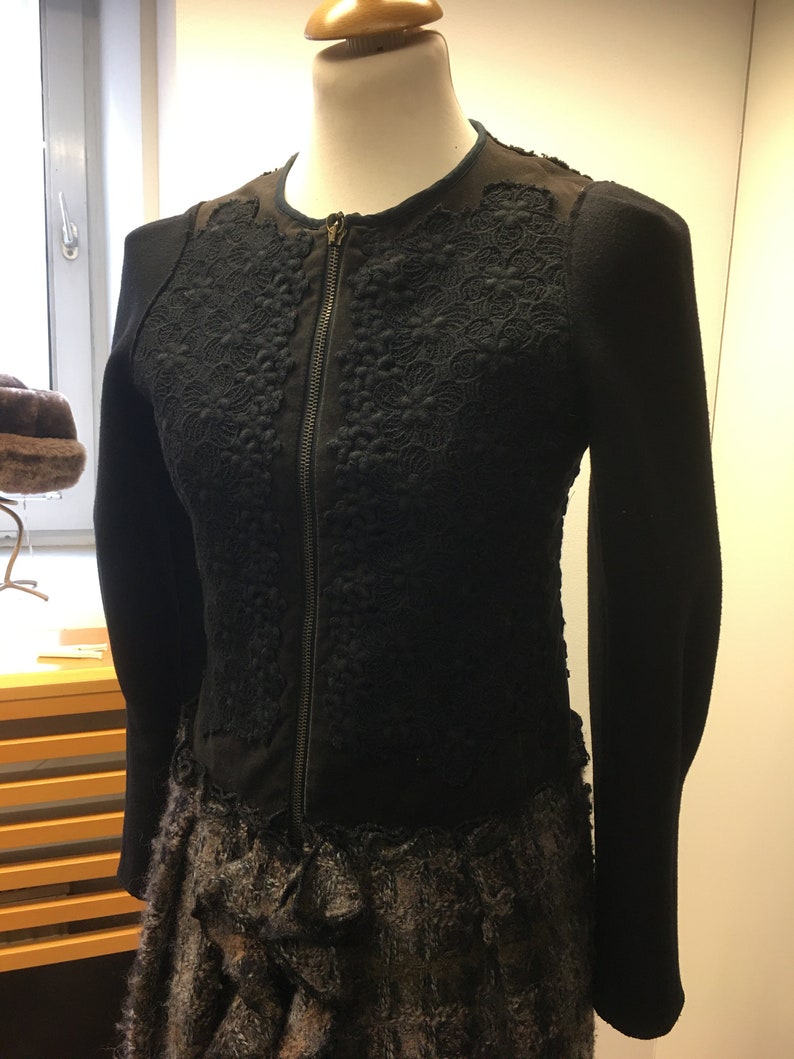 Puede incluir: Chaqueta negra con cierre de cremallera y detalles de encaje intrincados. La chaqueta tiene un dobladillo con volantes y est&aacute; hecha de una tela suave y texturizada.