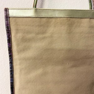 May include: A beige canvas tote bag with a gold leather top and a purple and green patterned trim.