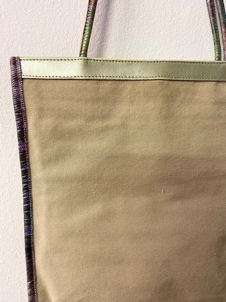 May include: A beige canvas tote bag with a gold leather top and a purple and green patterned trim.