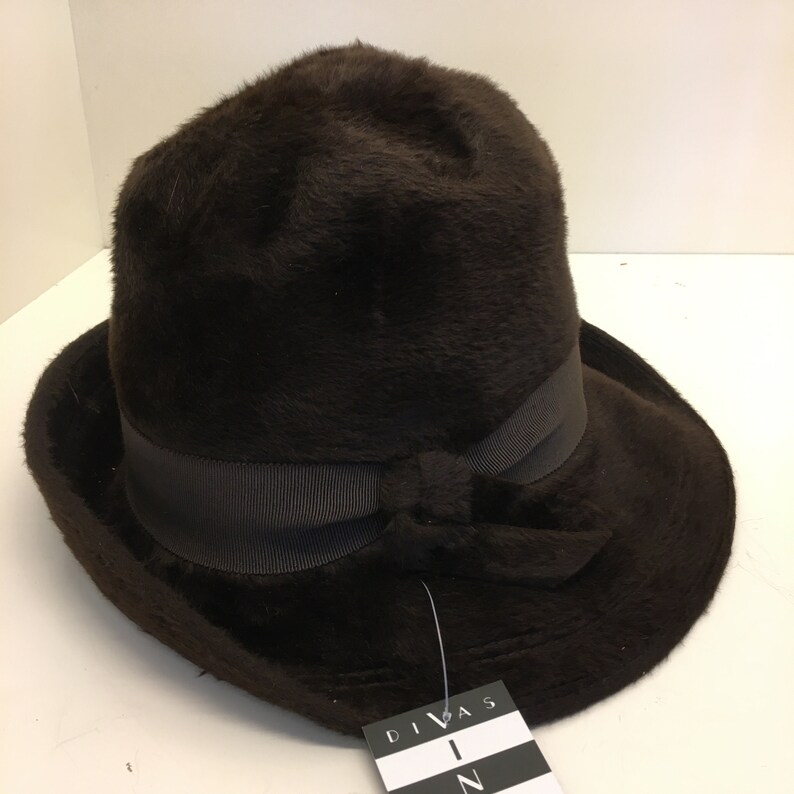 Puede incluir: Un sombrero de piel sint&eacute;tica marr&oacute;n con un ala ancha y una cinta negra atada en un lazo en la parte delantera. El sombrero tiene una etiqueta que dice "Divas In".