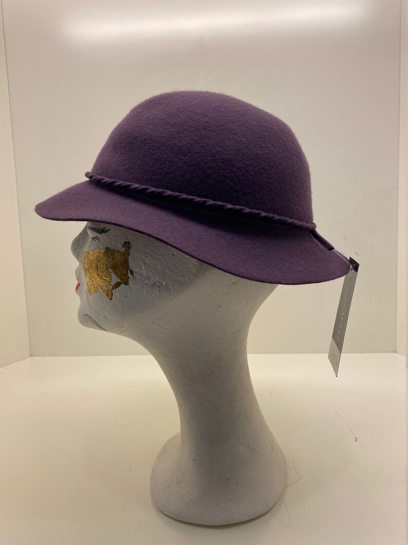 May include: A purple felt hat with a braided trim. The hat is on a white mannequin head.