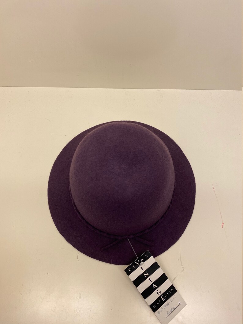 May include: A purple felt hat with a small brim and a bow on the front. The hat has a tag attached to it that says "Divas in Vintage Gallery".