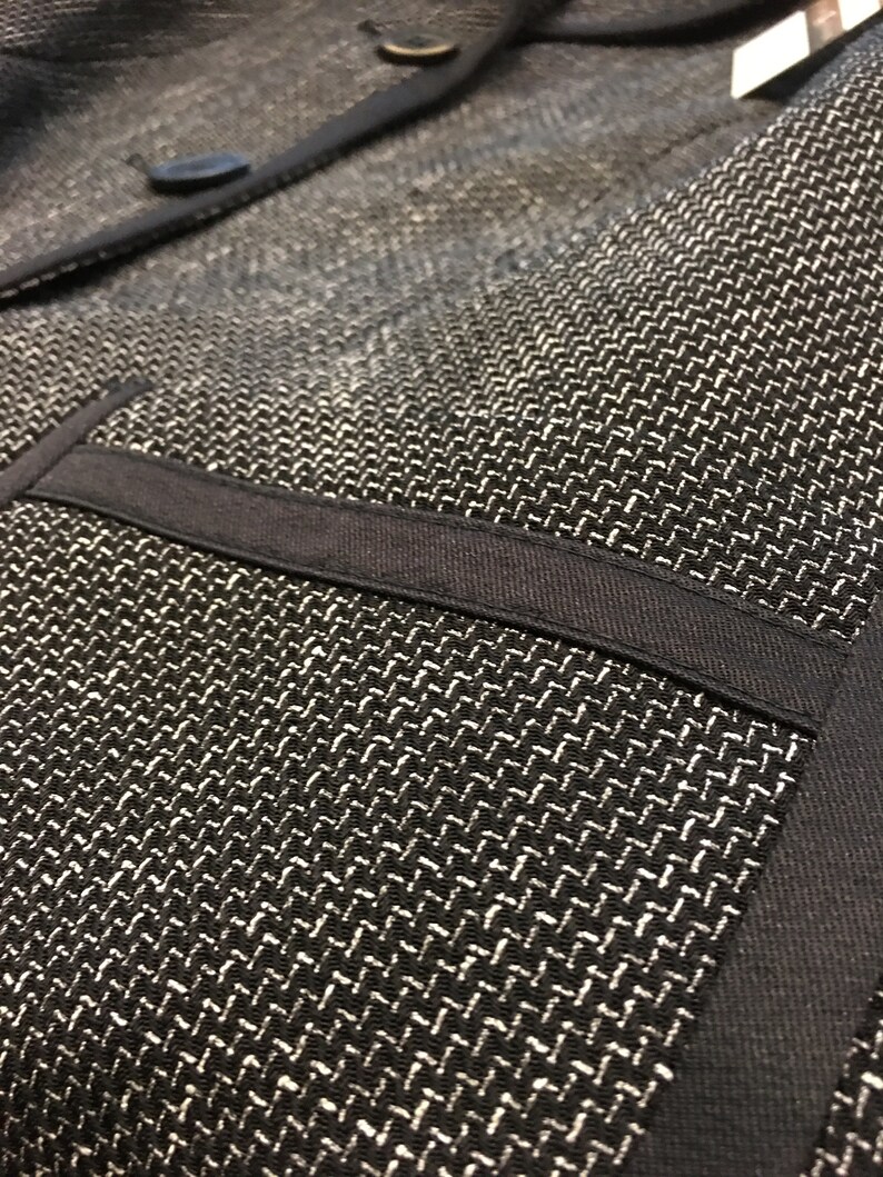 Puede incluir: Primer plano de una chaqueta negra con tejido texturizado. La chaqueta tiene un ribete negro y un bolsillo en la parte delantera.